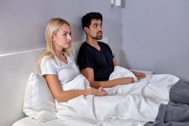 Couple watching tv in bed in cozy room at home looking at side, family concept. young caucasian man and woman in domestic clothes having rest, relaxing together. at weekends, holidays.