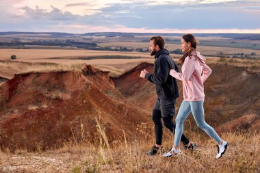 athletic male and female ogging along field outside the city at sunset, runners training in picturesque outdoor area. young caucasian couple in sportive clothes motivated and concentrated