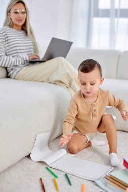 Little boy and mother spending weekends at home together, child drawing painting on floor while blonde mother working on laptop in the background. focus on cute child boy, indoors