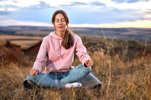 Young Female Keep Calm Sitting On Fitness Mat, Meditating Alone On Fresh Air. Attractive Young Lady In Sportive Leggins And Pink Hoodie relaxing, Having Rest. In Nature In The Morning