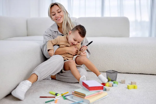 Cheerful young european mother play with excited smiling small son in living room at home. Overjoyed female have fun with little preschooler boy child, tickle giggle, enjoy family weekend together.