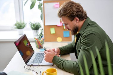 Young man sit using smartphone, having rest during work in bright modern office, alone. bearded caucasian guy in casual wear take a break typing message. business people, job, success