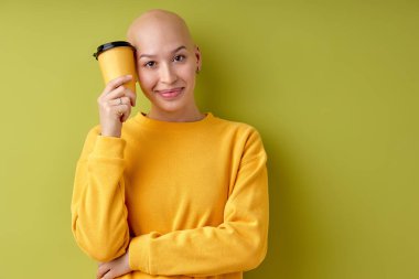 Closeup portrait of beautiful hairless woman on light green background. Smiling bald female looking at camera holding cup of coffee. Cheerful and satisfied young woman with shaved head.