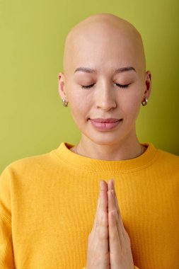 beautiful young caucasian bald hairless woman praying, keeping hands together, wearing casual clothes standing isolated over green background, asking for something, thanks God concept