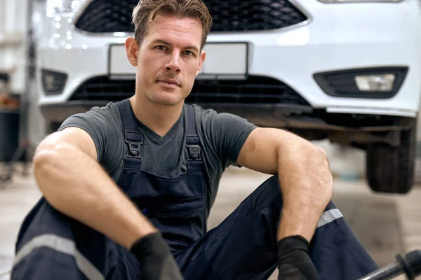 At auto service. Handsome young auto mechanic in uniform sit on floor having rest, looking at camera, after examining car. caucasian confident male in overalls and gloves take a break