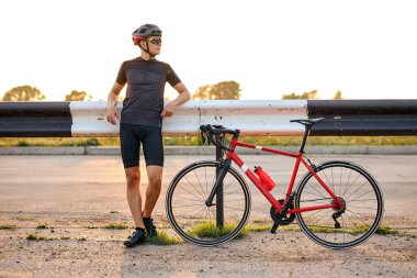 Cyclist in shorts and helmet standing on road in countryside having rest at summer season, confident caucasian guy in black sportswear taking a break, at sunny evening. sport, healthy lifestyle