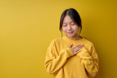 Thankful kind chinese female keeping hands on chest expressing gratitude, attractive asian lady in casual yellow shirt posing at camera with eyes closed. human emotions, people concept