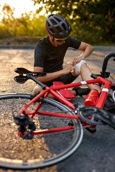 Accident man in sports concept. biker fall from bike and wrapping leg in bandage. injured young male bycyclist in black sportswear has an accident on bike, red bike on road next to him