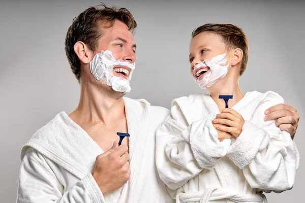 Happy dad and son with pleasant appearance, have shaving foam on faces, hold razors and going to shave, stand together, have fun, laughing. caucasian or american kid imitates father, isolated on gray