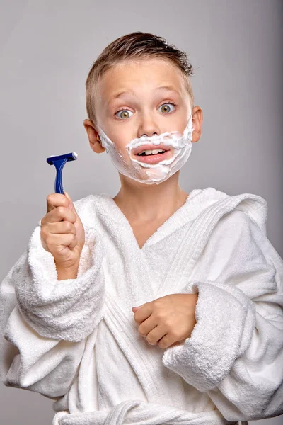 Surprised child boy son with pleasant appearance, have shaving foam on face, hold razor and going to shave, stand in frot of mirror isolated on gray background, young kid imitates father, in bathrobe