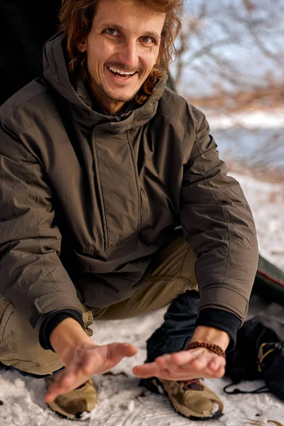 smiling man warm hands by fire, sit next to fire. Travel concept. happy guy enjoy exploring new places, camping. Burning wood at evening in nature, in winter forest, alone. portrait