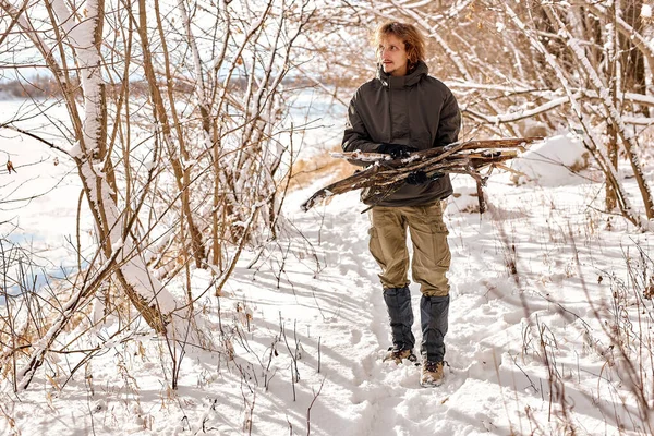 young caucasian male gathering wood for fire in forest, nature. lonely guy in warm clothes in snowy winter forest. bushcraft, camping, travel, active healthy lifestyle, nature concept.