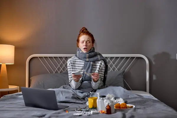 young red-haired sick girl holding smart phone sneezing, bless you. closeup portrait, laptop , pills, medicine antiviral antibiotics antipyretic painkillers are on bed