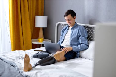 Yapay bacaklı engelli adam iş için dizüstü bilgisayar kullanıyor, evde uzaktan kumandalı bir işi var. Çevrimiçi iletişim kuran engelli genç beyaz adam. Yeni normal, sosyal uzaklık kavramı