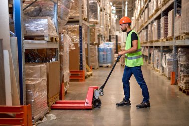 Genç erkek depo işçisi forklift kullanarak üretim deposuna teslimat yapıyor. Yelekli erkek personel ve iş yerinde turuncu kask. Forklift sürücüsü paletleri yüklüyor