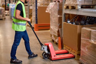 Depoda çalışan işçiler paletleri taşımak için el paleti istifleyicisi kullanırlar. İyi kargolar, iş yerindeki yeşil üniformalı tanımlanamayan erkek personel. Fabrika deposunda el arabası krikosu..