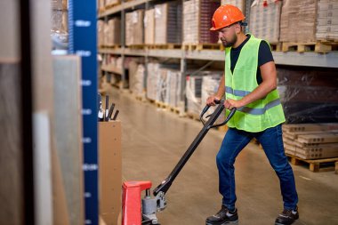 Genç erkek depo işçisi forklift kullanarak üretim deposuna teslimat yapıyor. Yelekli erkek personel ve iş yerinde turuncu kask. Forklift sürücüsü paletleri yüklüyor
