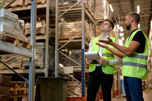 Retail Warehouse full of Shelves with Goods, Caucasian Male Workers Supervisors in working uniform, Holding Paper Tablet Discussing Product Delivery. In Distribution Logistics Center