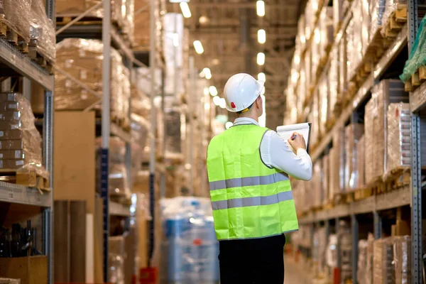 stock image rear view on young male in helmet with clipboard checking goods at warehouse, counting and controlling distribution. unrecognizable hardworking caucasian worker in green working uniform