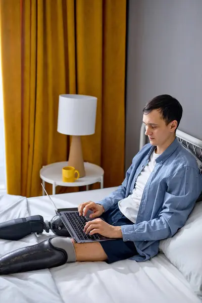 handicapped guy with artificial legs using laptop for work or studies, having online work at home. Disabled young Caucasian man communicating online. New normal, social distancing concept
