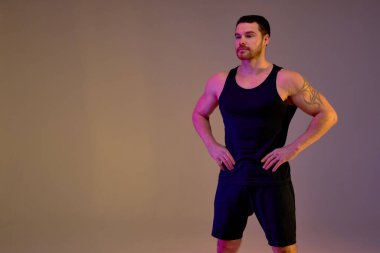 Stüdyoda elleri kalçasında dinlenen kaslı, güçlü sakallı bir adam, kolunda siyah spor kıyafetleriyle dövmesi olan genç bir sporcu öğrencileri çalıştırıyor, eğitmenlik yapıyor. Boşluğu kopyala