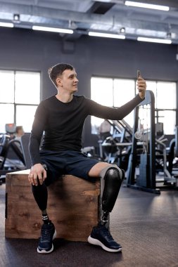 Sporcu görüntülü telefon görüşmesi yapıyor. Ahşap kutuda oturan, spor salonunda selfie çeken, uzun boy fotoğraf çeken bir adam. Spor merkezinde fotoğraf çeken engelli bir adam. Yaşam sevgisi. blogger çekim videosu