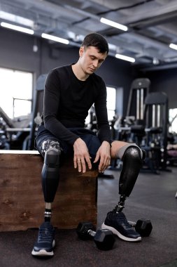 Sporcu yorgun, kutunun üzerinde oturuyor, yerdeki dambıllara bakıyor. Tam boy fotoğraf. Yorgunluk, fotoğrafın arka planında modern spor aletleri.