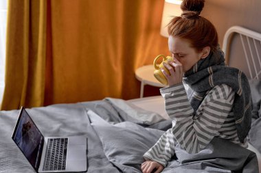 Kızıl saçlı bir kadın öksürüyor, hapşırıyor, internetten çalışırken kendini kötü hissediyor, kovid 19 sırasında serbest çalışıyor, koronavirüs, omikron. Baş ağrısı olan bir kadın sıcaklığı düşürmek için sıcak çay içiyor.