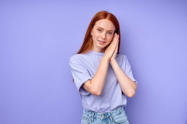 Gülümseyen kızıl saçlı kadın, uyku jesti gösteriyor, katlanmış ellerle kameraya bakıyor. Neşeli pozitif model uykulu işareti güldürüyor yakın plan portresi izole edilmiş mavi arka plan yatağı