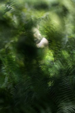 Omuzlarının üzerinde kısmen aydınlanmış dantel kanatlı genç bir kadın, rüya gibi yeşil bitki dokularında eğreltiotu tutuyor..