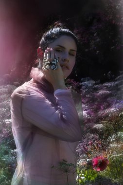 Şeffaf bluz giymiş genç bir bayan, yanağında bir Delias Diaphana kelebeği tutarken, bir Laceflower tarlasında renklendirme efektiyle ayakta duruyor..