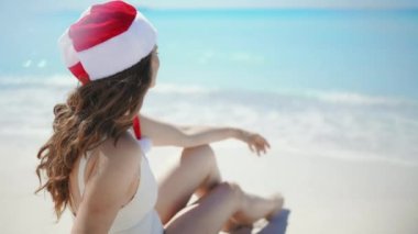 Beyaz mayo giyen, uzun dalgalı saçlı ve çizgili Noel şapkalı bir kadının kumsalda oturduğunu gördüm..