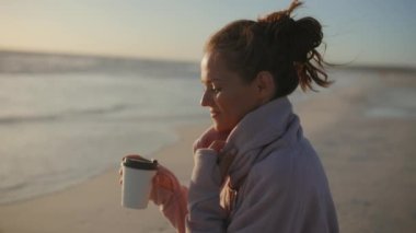 gülümseyen, orta yaşlı, rahat kazaklı bir kadın ve kumsalda bir fincan sıcak çikolata..