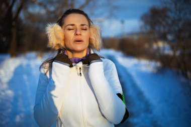 Beyaz ceketli sağlıklı bir kadın, kulak ısıtıcı ile kışın şehir parkında egzersiz yaptıktan sonra soluklanıyor..