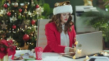 Noel zamanı. Gülümseyen orta yaşlı, Noel Baba şapkalı, kırmızı ceketli, modern yeşil ofiste Noel ağacıyla çalışan küçük işletme sahibi kadının portresi..