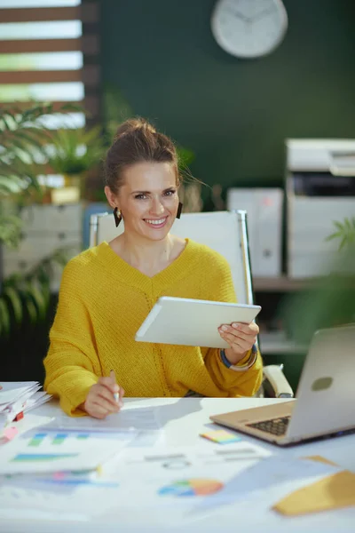 在现代的绿色办公室里 穿着黄色毛衣 头戴平板电脑和笔记本电脑的快乐的中年小企业主女人 — 图库照片