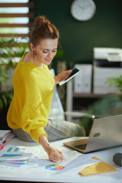 在现代的绿色办公室里 穿着黄色毛衣 使用平板电脑和文件的快乐的小企业主女人 — 图库照片