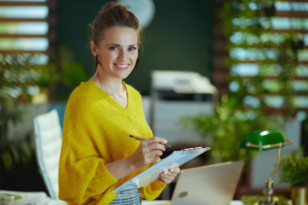 幸せなエレガントな中年中小企業所有者女性で黄色セーターでクリップボードでザ現代緑のオフィス — ストック写真