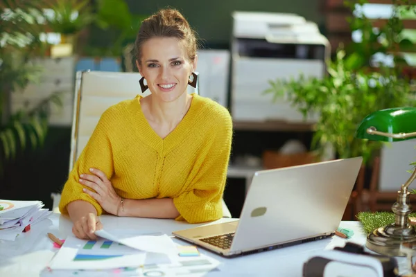 幸せなエレガントな40歳の小さなビジネスの所有者女性で黄色のセーターとノートパソコン作業のドキュメントで現代の緑のオフィス — ストック写真