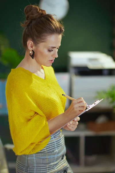 スタイリッシュな40歳の小さなビジネスの所有者女性で黄色のセーターでクリップボードで現代の緑のオフィス — ストック写真