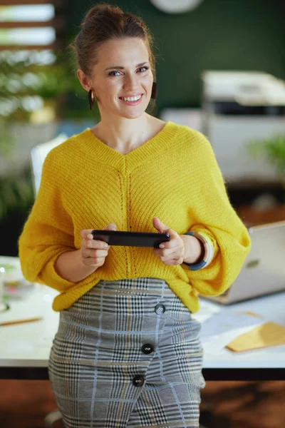 幸せなトレンディーな中年中小企業所有者女性で黄色セーターとともにスマートフォンで現代緑のオフィス — ストック写真