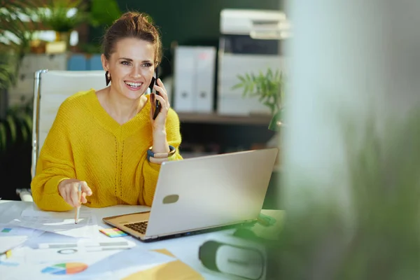 笑顔トレンディーな中年中小企業のオーナー女性で黄色のセーターでラップトップはスマートフォンで話すと現代の緑のオフィスで文書を扱う — ストック写真