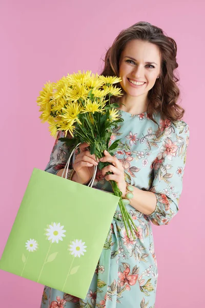 エレガントな40歳の女性とともに長い波状ブルネットの髪とともに黄色の菊の花と緑のショッピングバッグにピンクの背景 — ストック写真