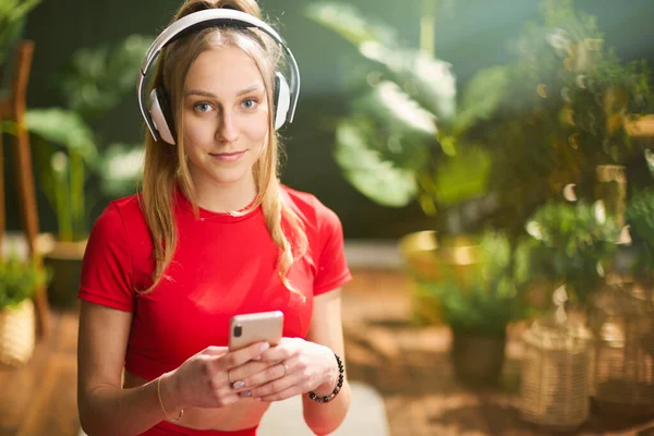現代の緑のリビングルームでスマートフォンを使用してヘッドフォンと赤フィットネス服の現代女性の肖像画 — ストック写真