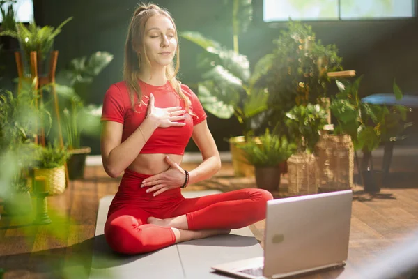 modern woman in red fitness clothes with yoga mat and laptop doing yoga in the modern green living room.