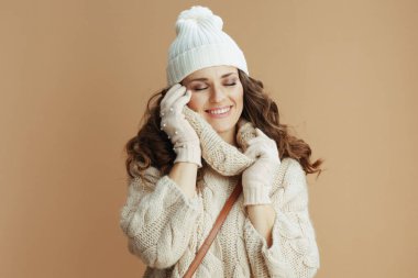 Merhaba kış. Bej kazak, eldiven ve bej arka planda şapka giyen 40 yaşında rahat bir kadın..