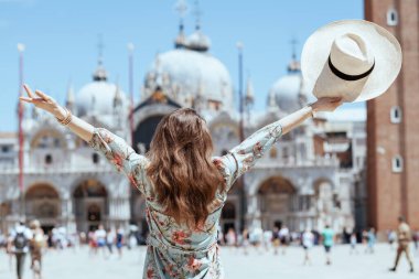 Seen from behind traveller woman in floral dress with hat at San Marco square in Venice, Italy with raised arms rejoicing. clipart