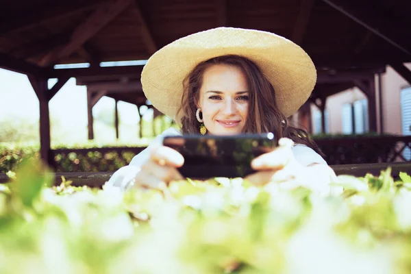 白いシャツを着た現代的な中年女性の笑顔と牧場でスマートフォンを使ってテキストメッセージを送る帽子 — ストック写真