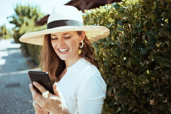 トレンディーな笑顔40歳の女性の白いシャツに帽子緑の壁の近くの屋外でスマートフォンアプリを使用して — ストック写真