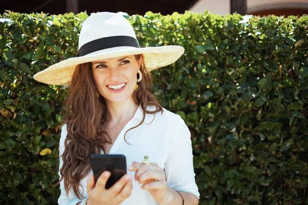緑の壁の近くの屋外でスマートフォンアプリを使用して帽子と白いシャツの幸せなエレガントな主婦 — ストック写真